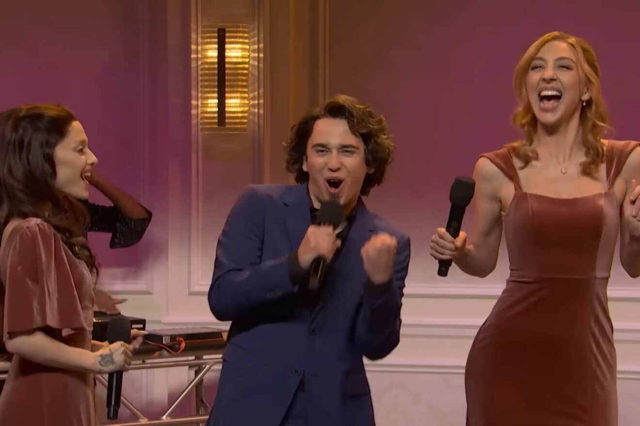 A man with medium-toned skin, wearing a suit, dances between two women in mauve dresses, each with light-toned skin.